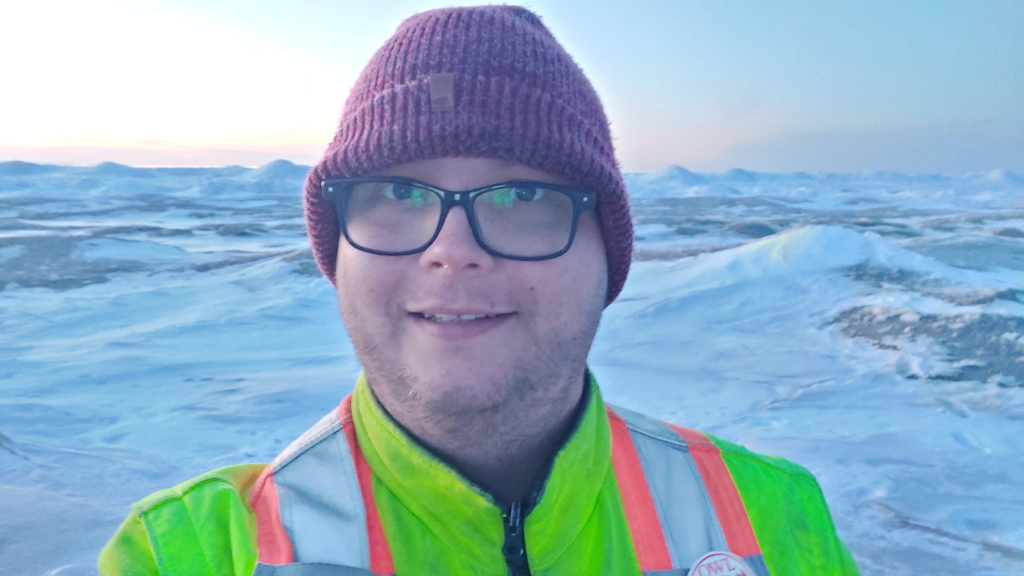 A man wears a red toque and yellow high visibility winter coat, standing in front of a landscape that looks like frozen waves